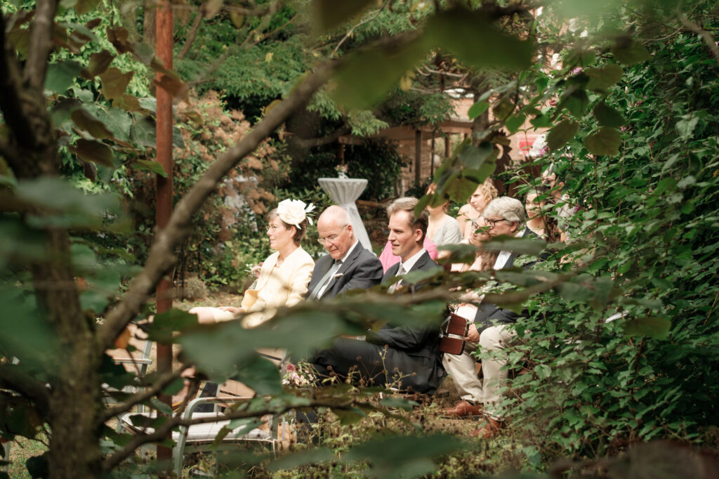 Blick durch die Bäume auf die Trauung im Garten, Hochzeit Anne & Phil auf dem Hof Gottschalk in Helpsen