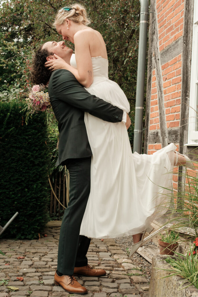 Bräutigam hebt die Braut am Fachwerkhaus hoch, Hochzeit Anne & Phil auf dem Hof Gottschalk in Helpsen